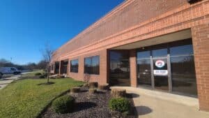 Single-story brick office building with several windows and glass double doors, surrounded by grass, small shrubs, and a clear blue sky.