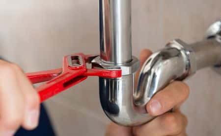 Close-up of hands using a red pipe wrench to tighten a metal pipe joint under a sink.