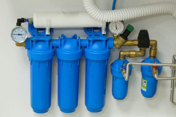 Three-stage water filtration installation with blue filter housings, pressure gauges, and connected pipes and hoses, neatly mounted on a wall.