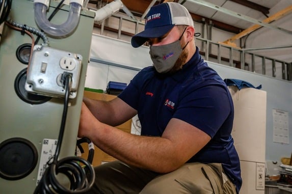 A service technician is in a home basement, wearing a mask, working on a heater.