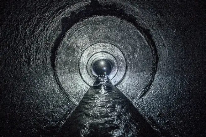 A dark, circular tunnel with water flowing through the center, illuminated by a light source at the far end—ideal for showcasing trenchless sewer line repair methods.