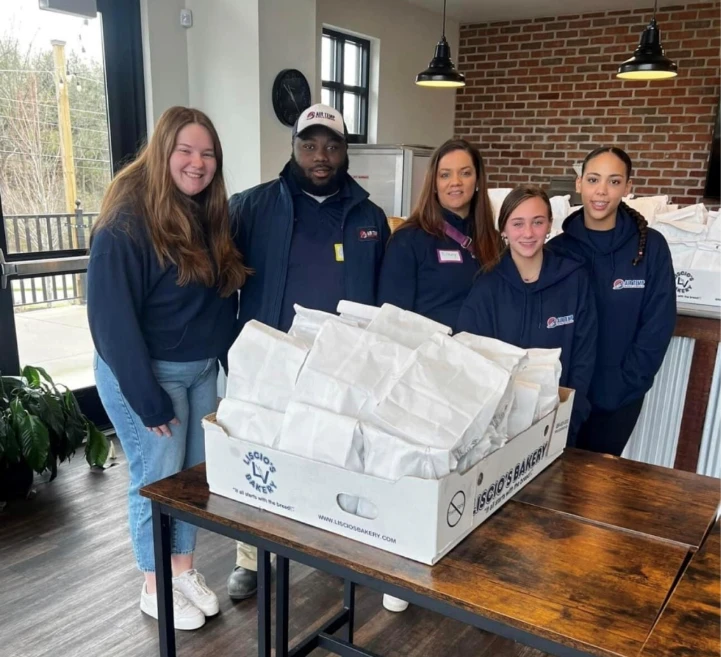Five people in matching navy hoodies stand behind a table with a large box filled with white paper bags, indoors by a brick wall and window.