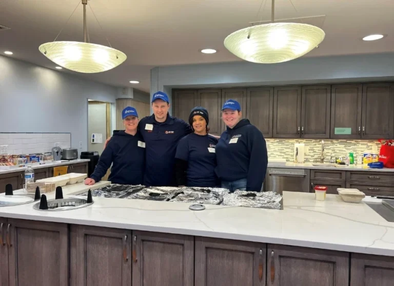 Four people in matching uniforms and hats stand behind a kitchen counter covered with foil trays in a modern kitchen setting.