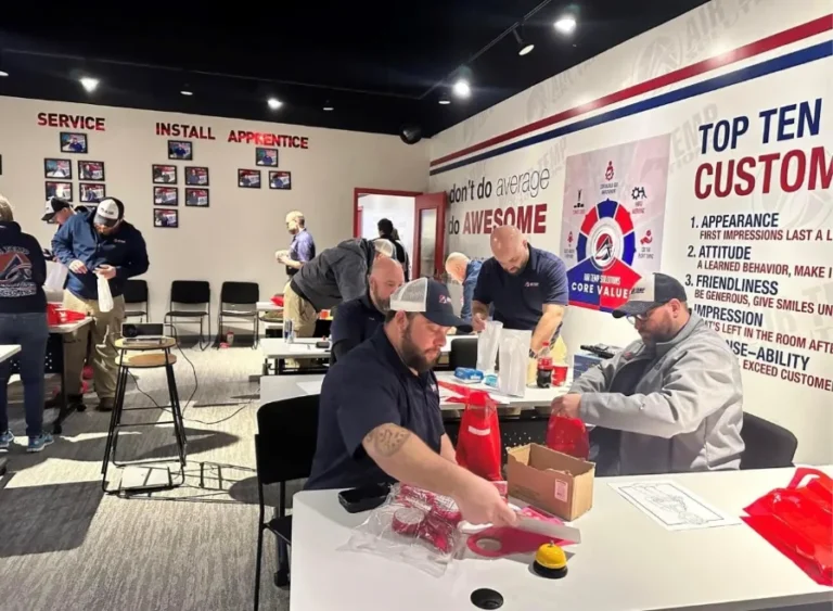 Several people in work uniforms assemble items at tables in a brightly lit room with company values and customer service posters on the walls.