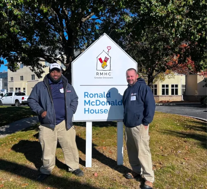 Two men in jackets stand in front of a Ronald McDonald House sign on a sunny day, with trees and a building in the background.