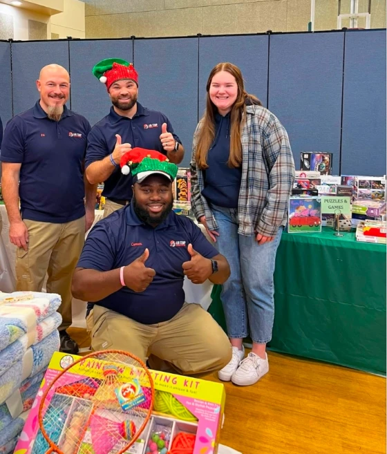 Four people stand and kneel by a table with toys, games, and blankets. Two men wear elf hats. All are smiling indoors at what appears to be a donation or holiday event.