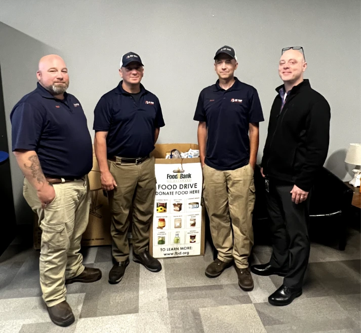 Four men stand beside a food donation box labeled "Food Bank Food Drive" filled with food items, in an indoor setting with gray walls and tiled floor.